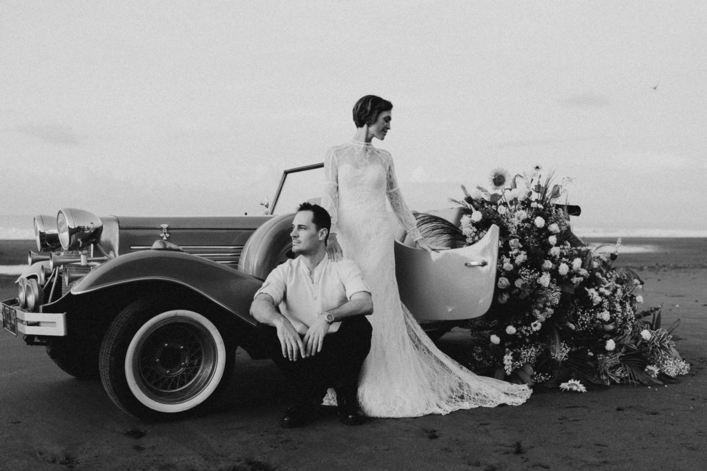 black and white image of bride and groom standing next to a vintage car on the beach at their Bali elopement