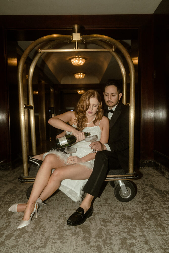 Couple celebrating their engagement with champagne while sitting on a luggage cart at The Benson Hotel for their Portland engagement photos.